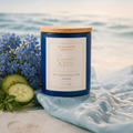 Sueño Azul soy candle with lid on, surrounded by bluebells and cucumber leaves in a dreamy coastal scene